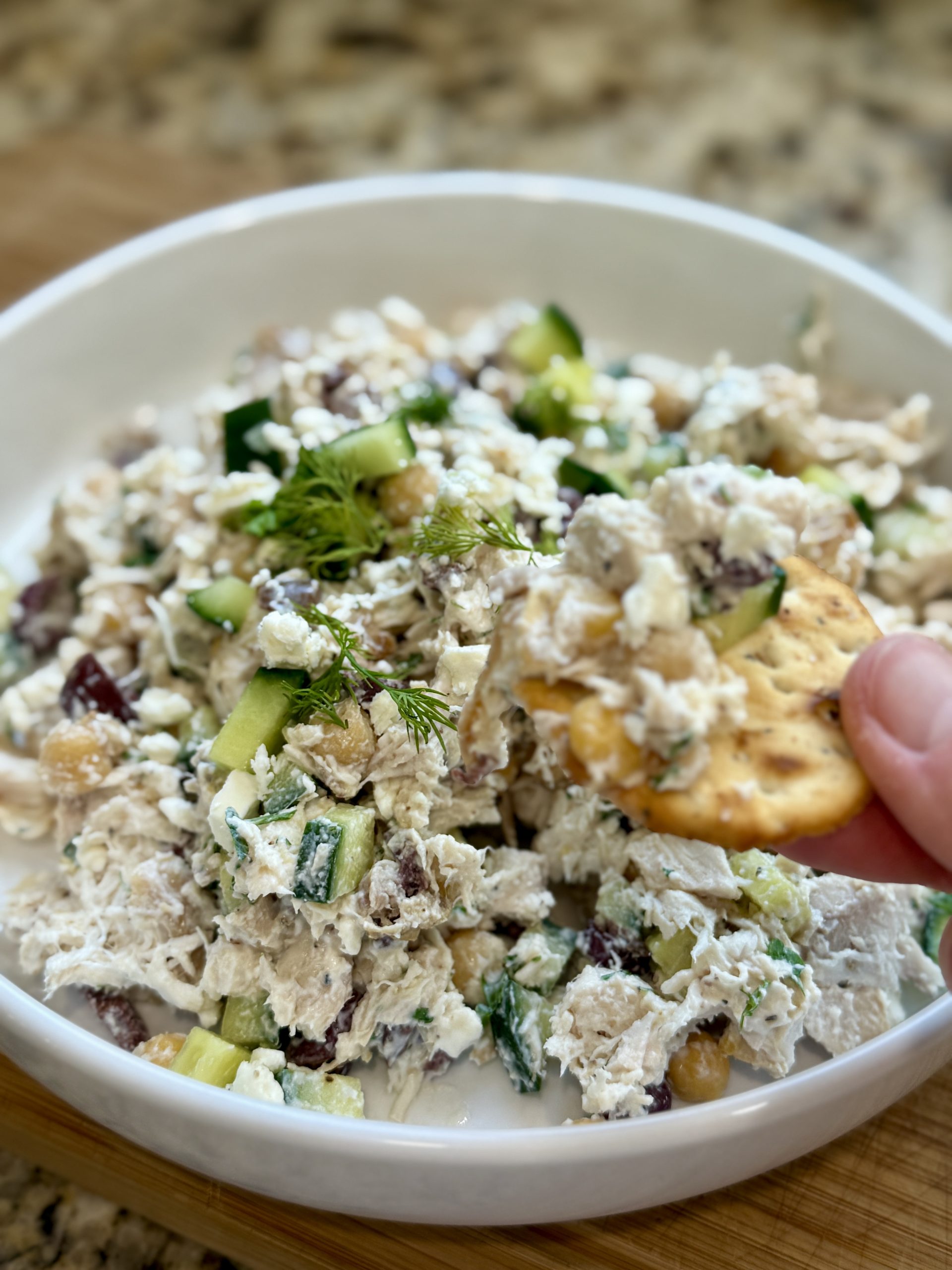 A close-up of a bowl of chicken salad with cucumbers, dill, and grapes, being scooped up with a round cracker by a hand. The background shows a wooden surface and a granite countertop.