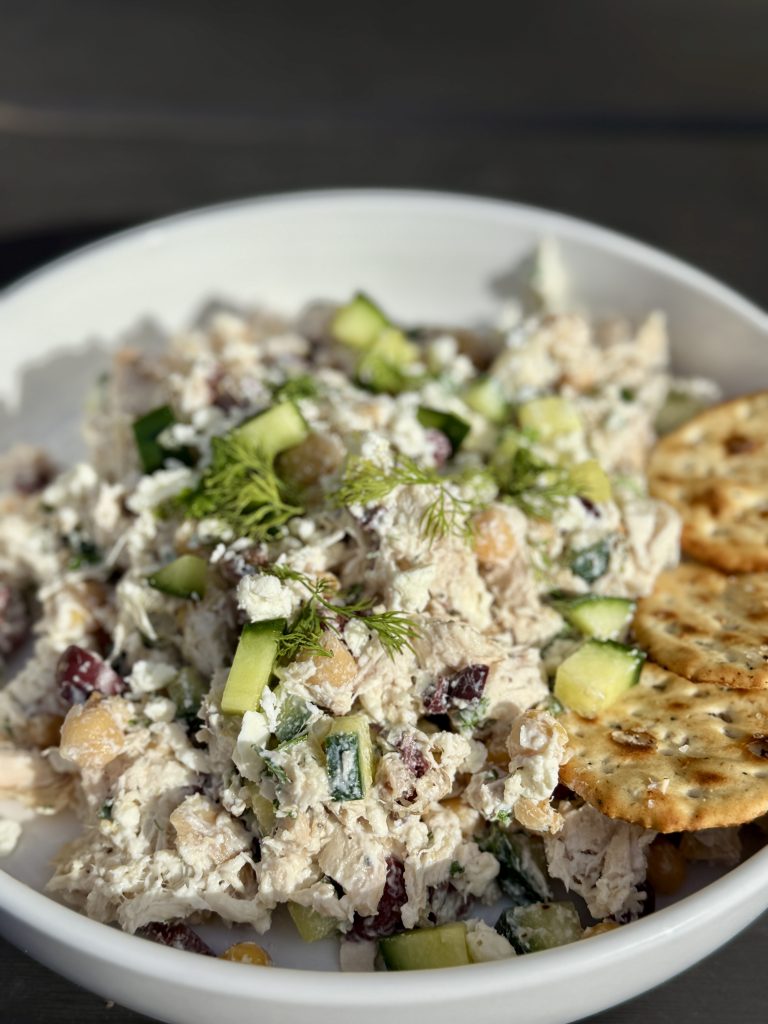 A white bowl filled with chicken salad, garnished with cucumber, dill, chickpeas, cranberries, and feta cheese, served with several round crackers on the side.