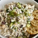 A bowl filled with a chicken salad mixed with cucumbers, chickpeas, olives, and herbs, garnished with fresh dill, and served with several round crackers on the side.