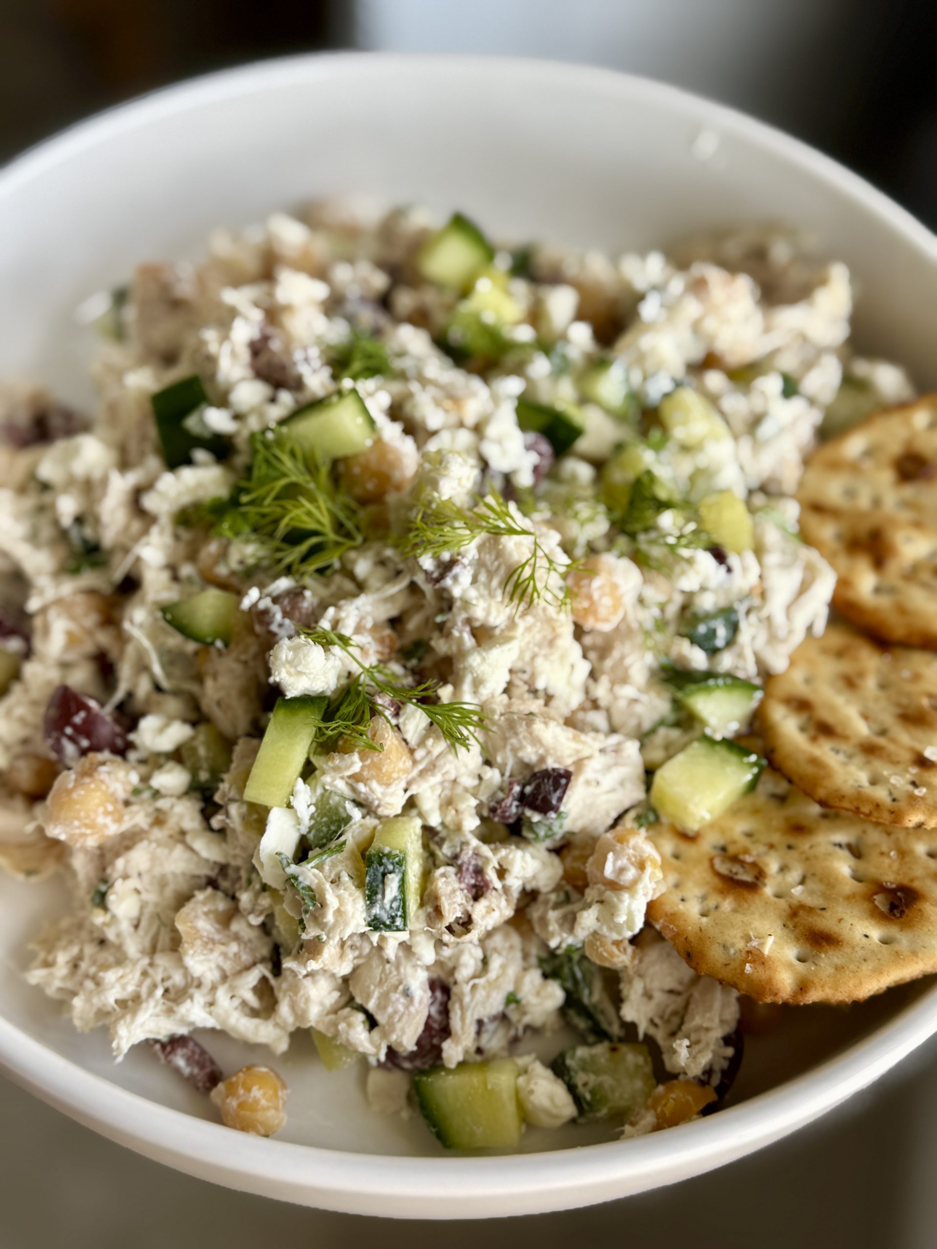 A bowl filled with a chicken salad mixed with cucumbers, chickpeas, olives, and herbs, garnished with fresh dill, and served with several round crackers on the side.