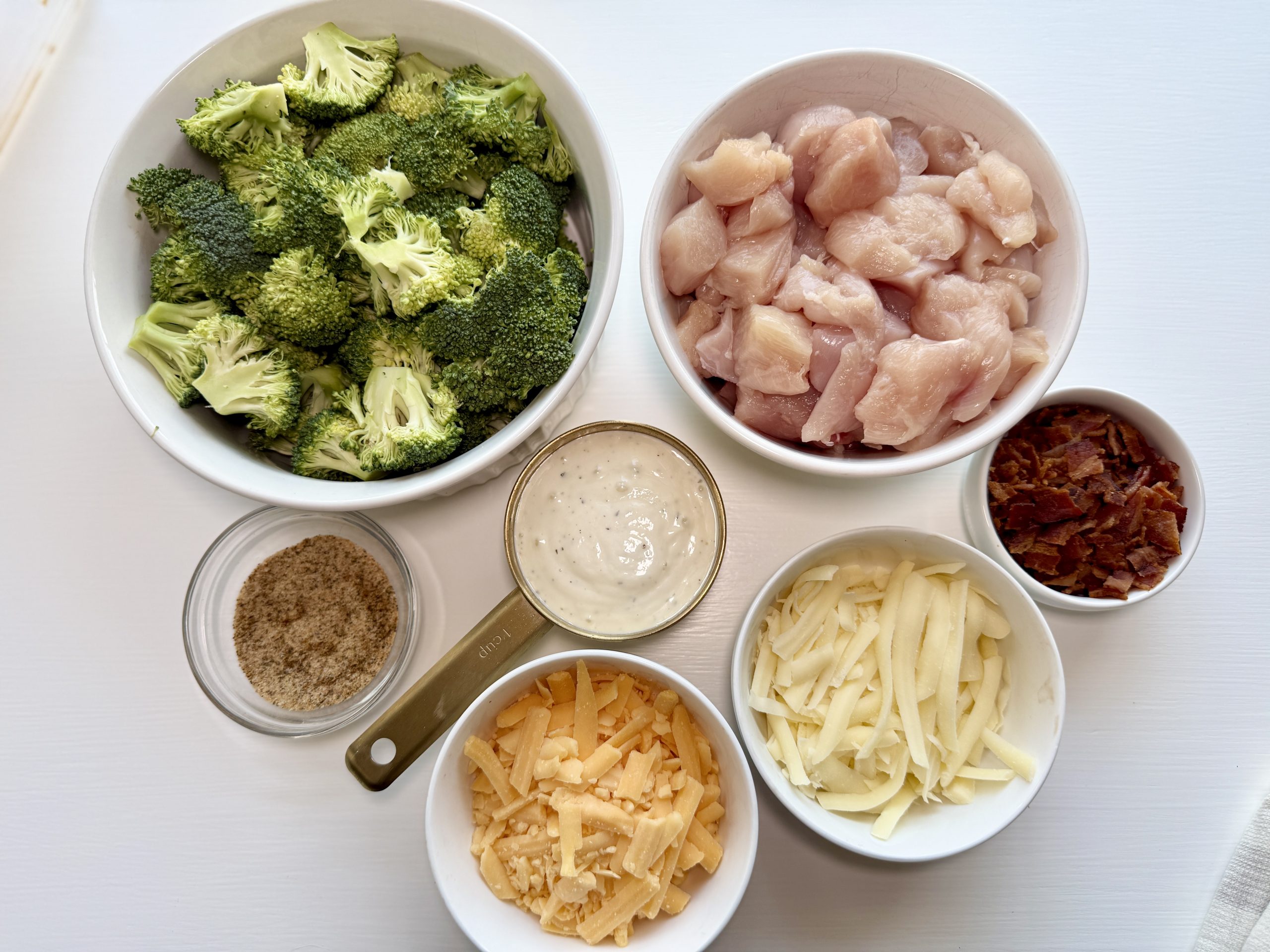 Bowls of raw broccoli florets, diced raw chicken, shredded cheddar and mozzarella cheeses, bacon bits, ranch dressing, and a measuring cup of seasoning on a white surface.
