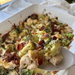 A close-up of a serving spoon lifting a portion of a baked casserole with broccoli, chicken, bacon, and melted cheese from a white baking dish near a window.