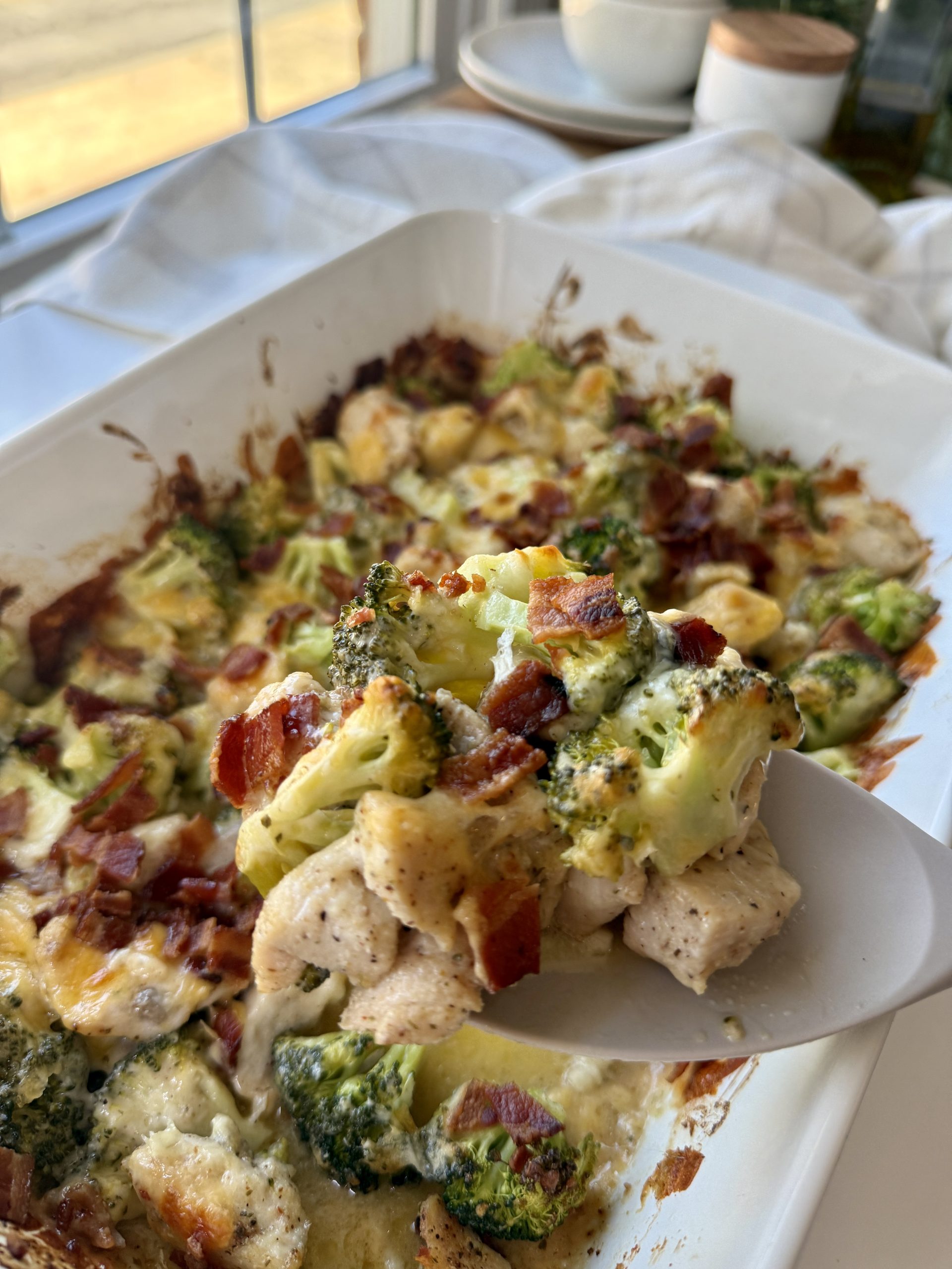 A close-up of a serving spoon lifting a portion of a baked casserole with broccoli, chicken, bacon, and melted cheese from a white baking dish near a window.