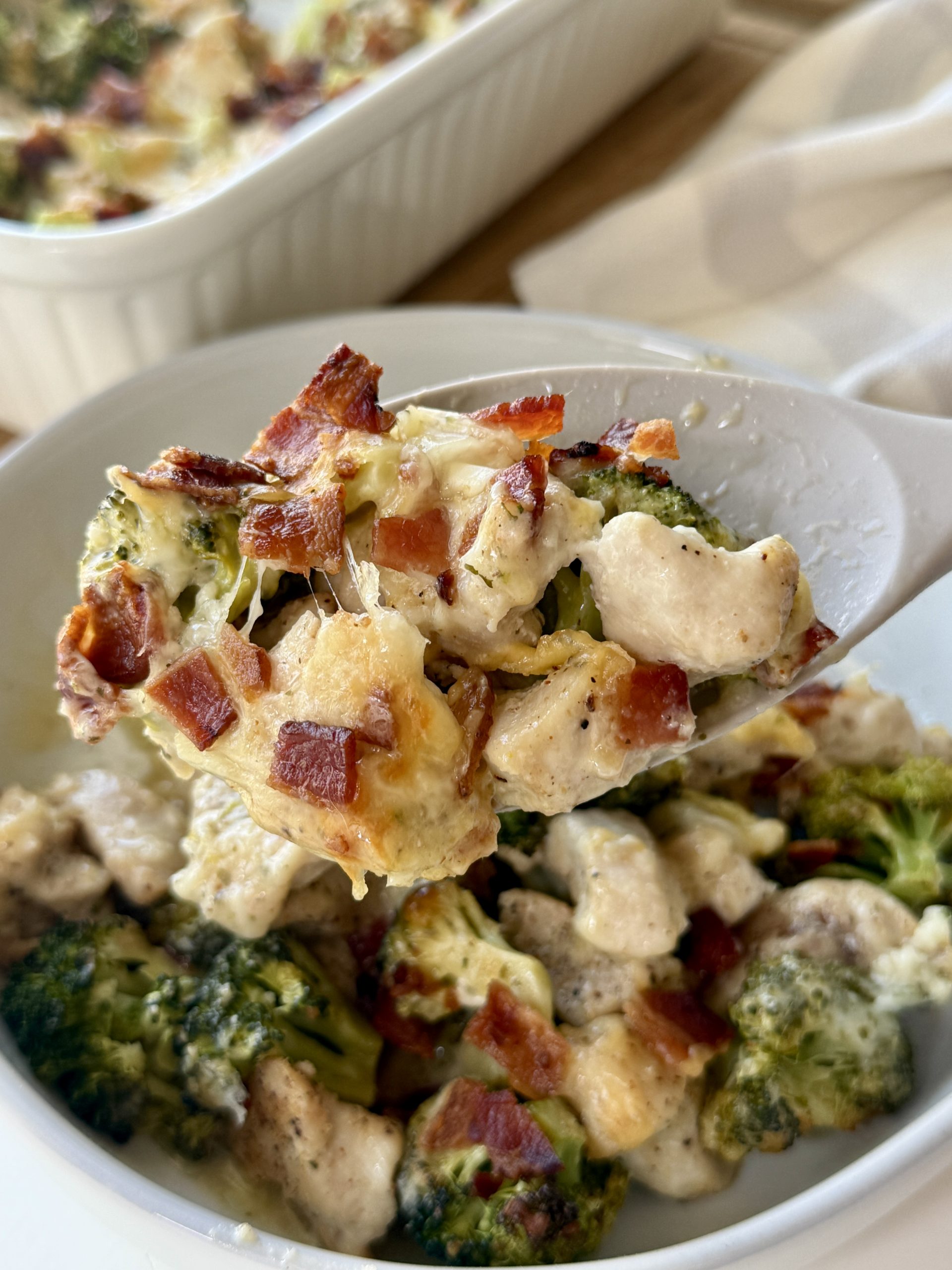 A close-up of a serving spoon holding a portion of creamy chicken and broccoli casserole topped with crispy bacon pieces, with the dish in a white baking dish in the background.