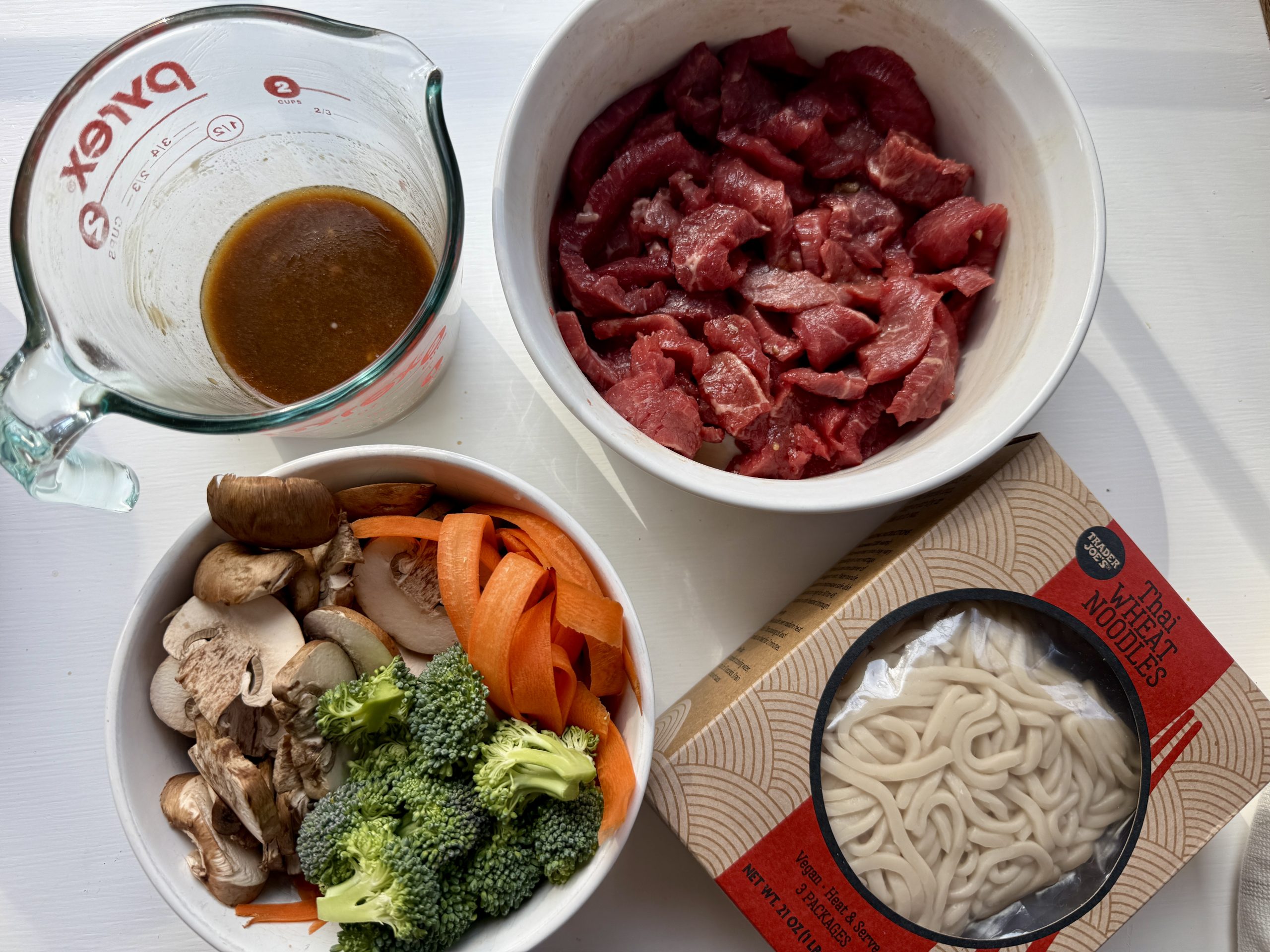 Four bowls on a white surface: sliced raw beef, brown sauce in a Pyrex cup, assorted vegetables (broccoli, mushrooms, carrots), and a pack of udon noodles.