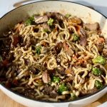 A pan filled with stir-fried noodles, beef slices, broccoli, carrots, and other vegetables, garnished with sesame seeds.