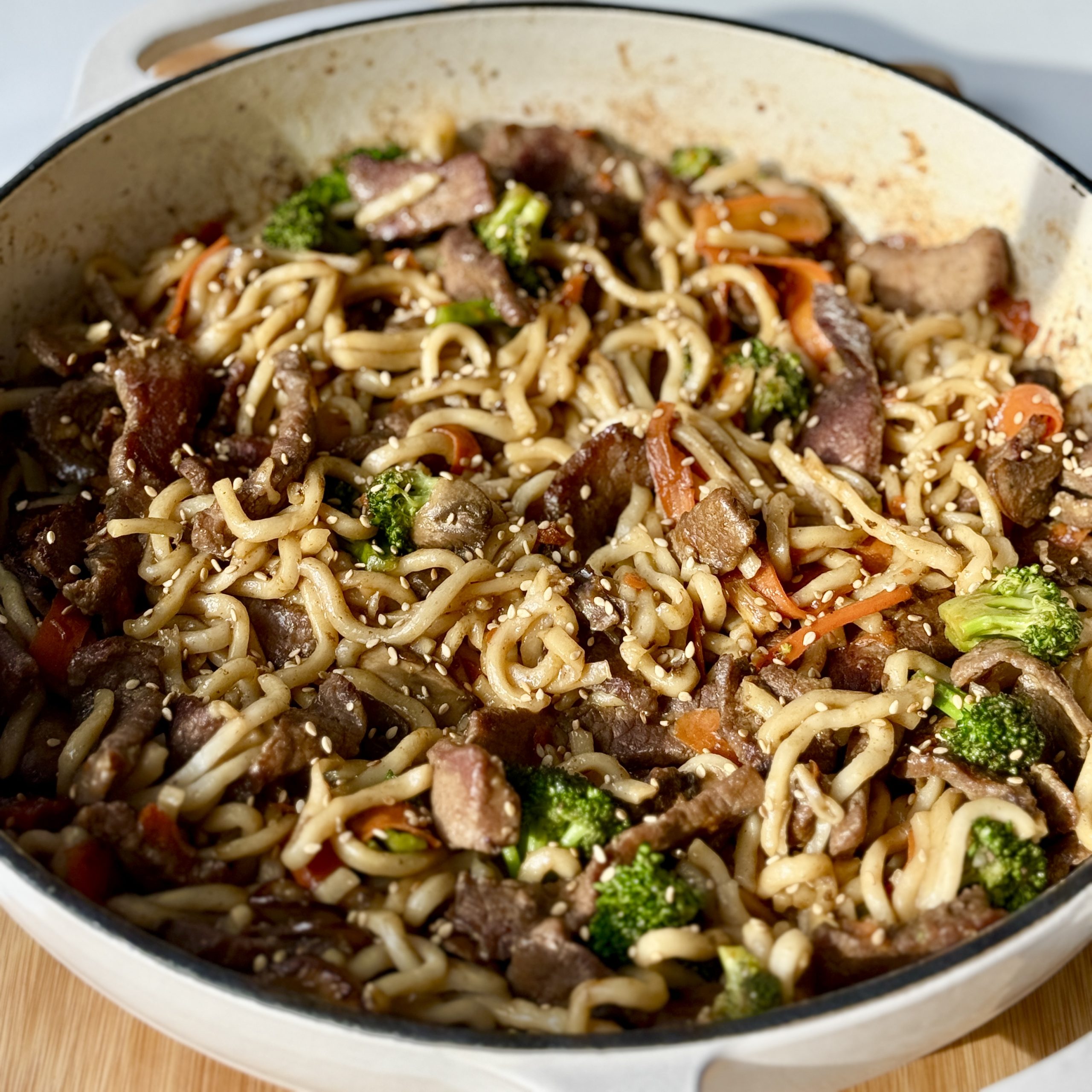 A pan filled with stir-fried noodles, beef slices, broccoli, carrots, and other vegetables, garnished with sesame seeds.