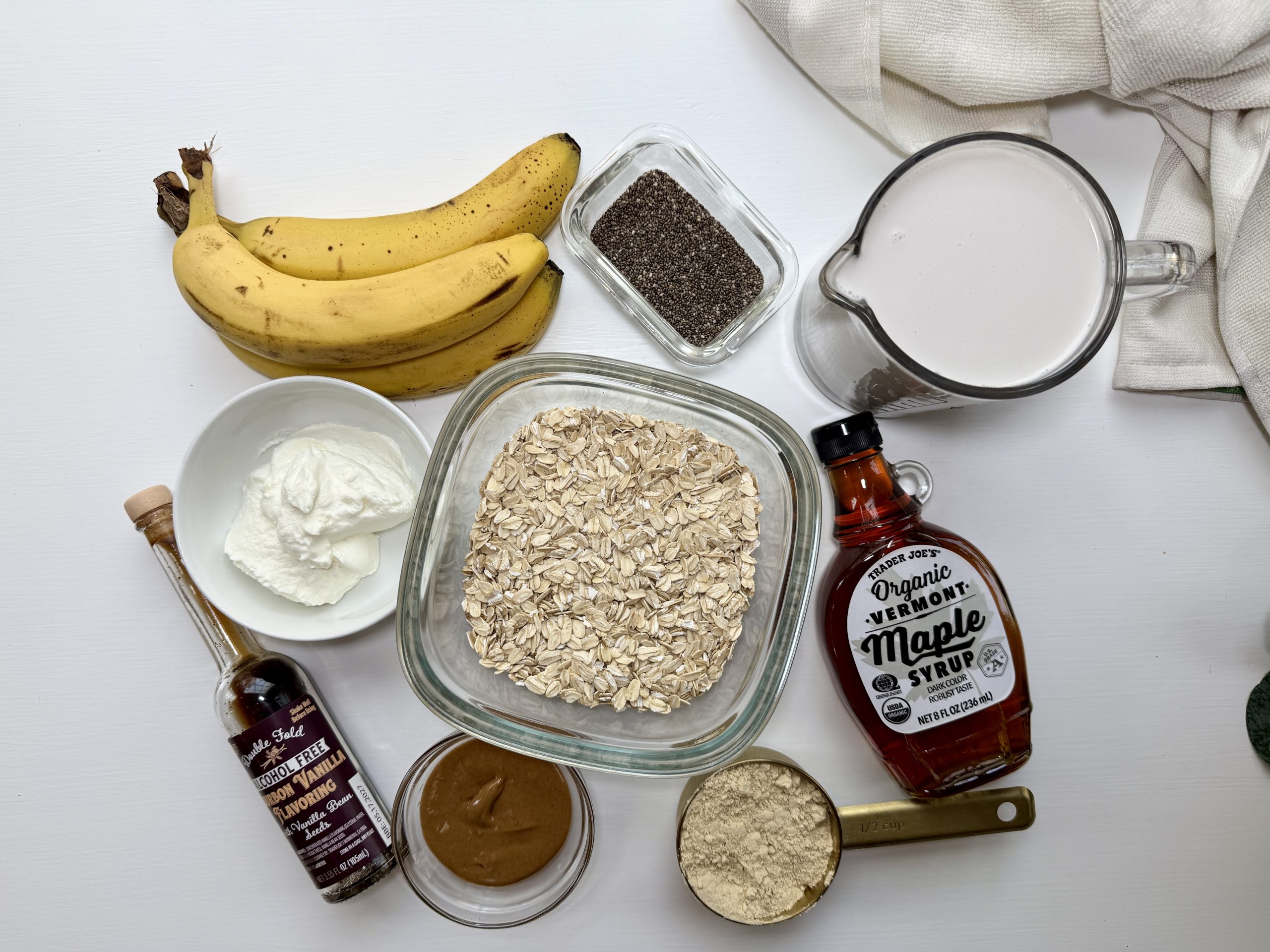 Overhead view of ingredients on a white surface: bananas, chia seeds, milk, maple syrup, rolled oats, peanut butter, Greek yogurt, vanilla extract, and protein powder. White towel in the top right corner.