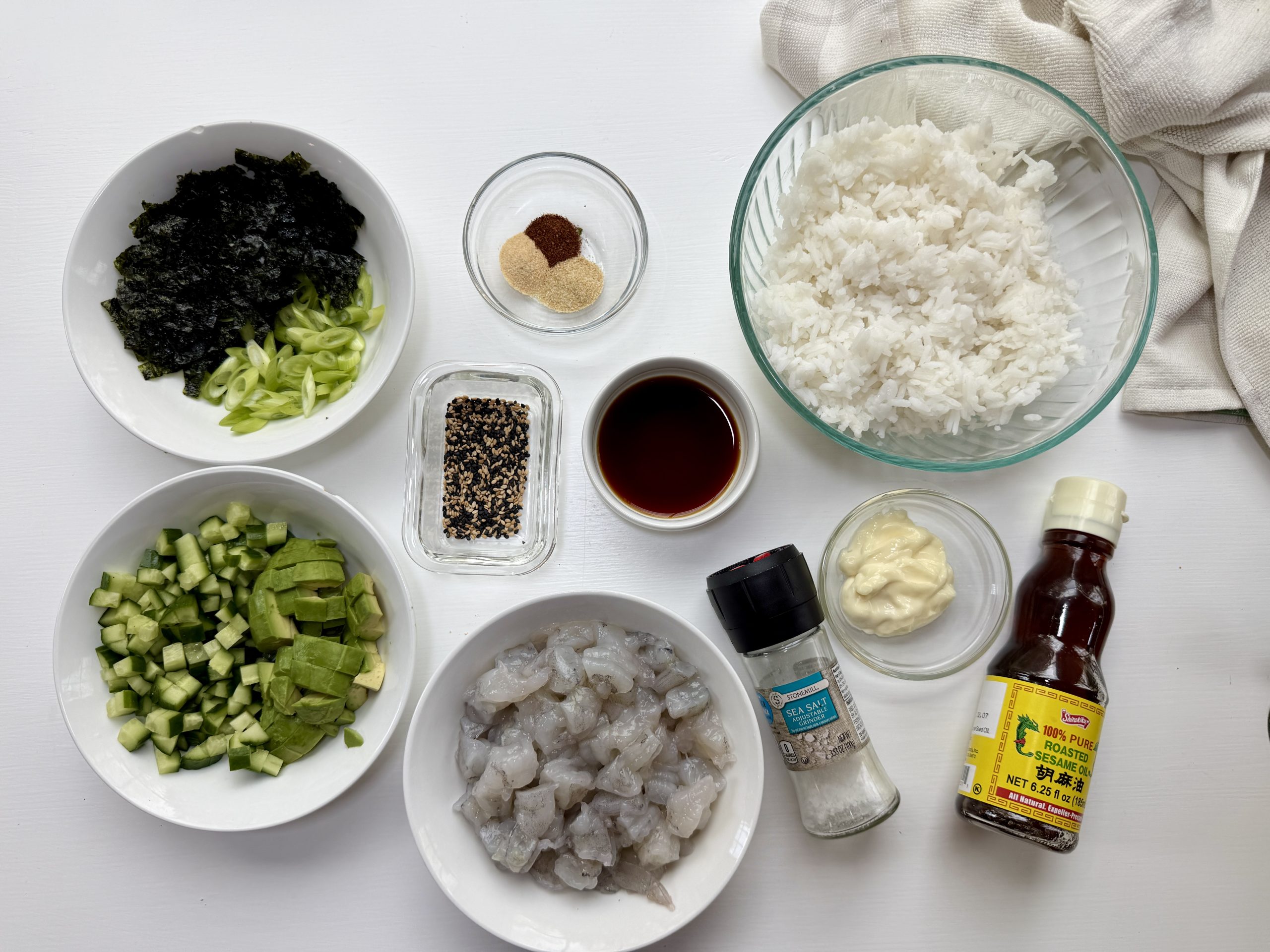 A flat lay of ingredients in bowls on a white surface: cooked rice, soy sauce, mayonnaise, bottle of spicy mayo, shrimp, diced cucumber, sliced green onion, seaweed, sesame seeds, seasonings, and salt.