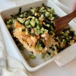A close-up of a baked sushi dish topped with chopped cucumbers, seaweed strips, and pieces of baked fish or seafood, served in a white square baking dish with a wooden spatula lifting a portion.
