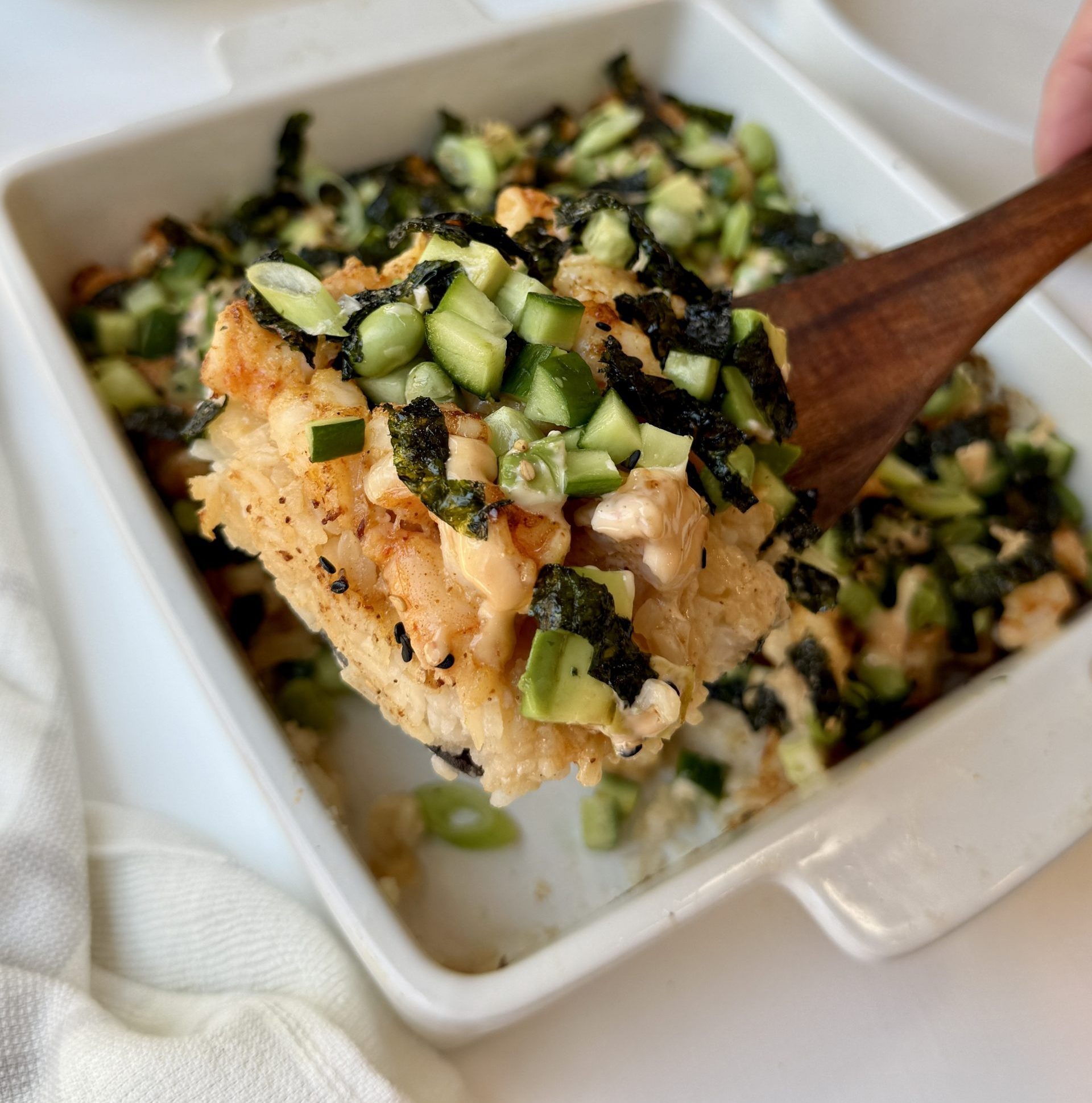A close-up of a baked sushi dish topped with chopped cucumbers, seaweed strips, and pieces of baked fish or seafood, served in a white square baking dish with a wooden spatula lifting a portion.