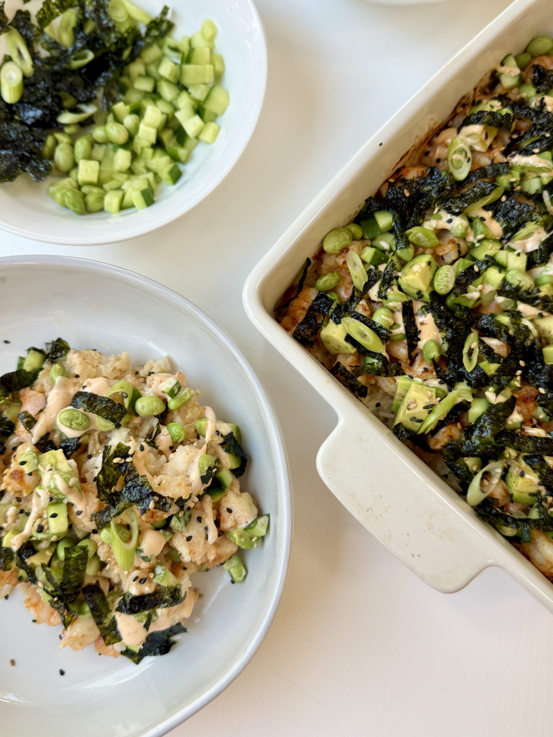 A close-up of a baked sushi dish topped with chopped cucumbers, seaweed strips, and pieces of baked fish or seafood, served in a white square baking dish with a wooden spatula lifting a portion.