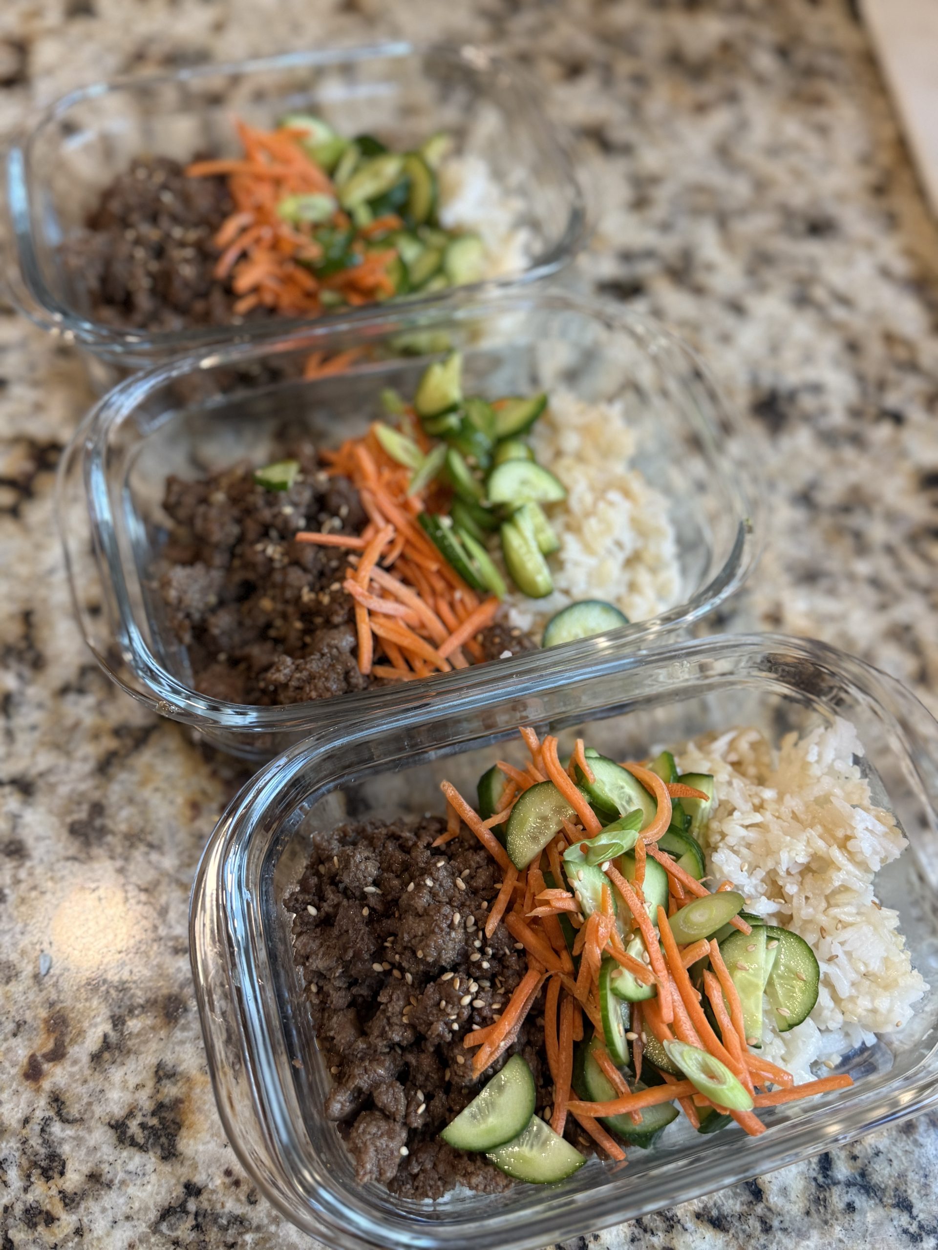 Three glass meal prep containers are filled with cooked ground beef, sliced cucumbers, shredded carrots, and white rice, arranged neatly on a speckled granite countertop.