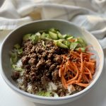 A bowl of white rice topped with cooked ground beef, sliced green onions, julienned carrots, and sliced cucumbers, placed on a white surface with a beige and white cloth in the background.