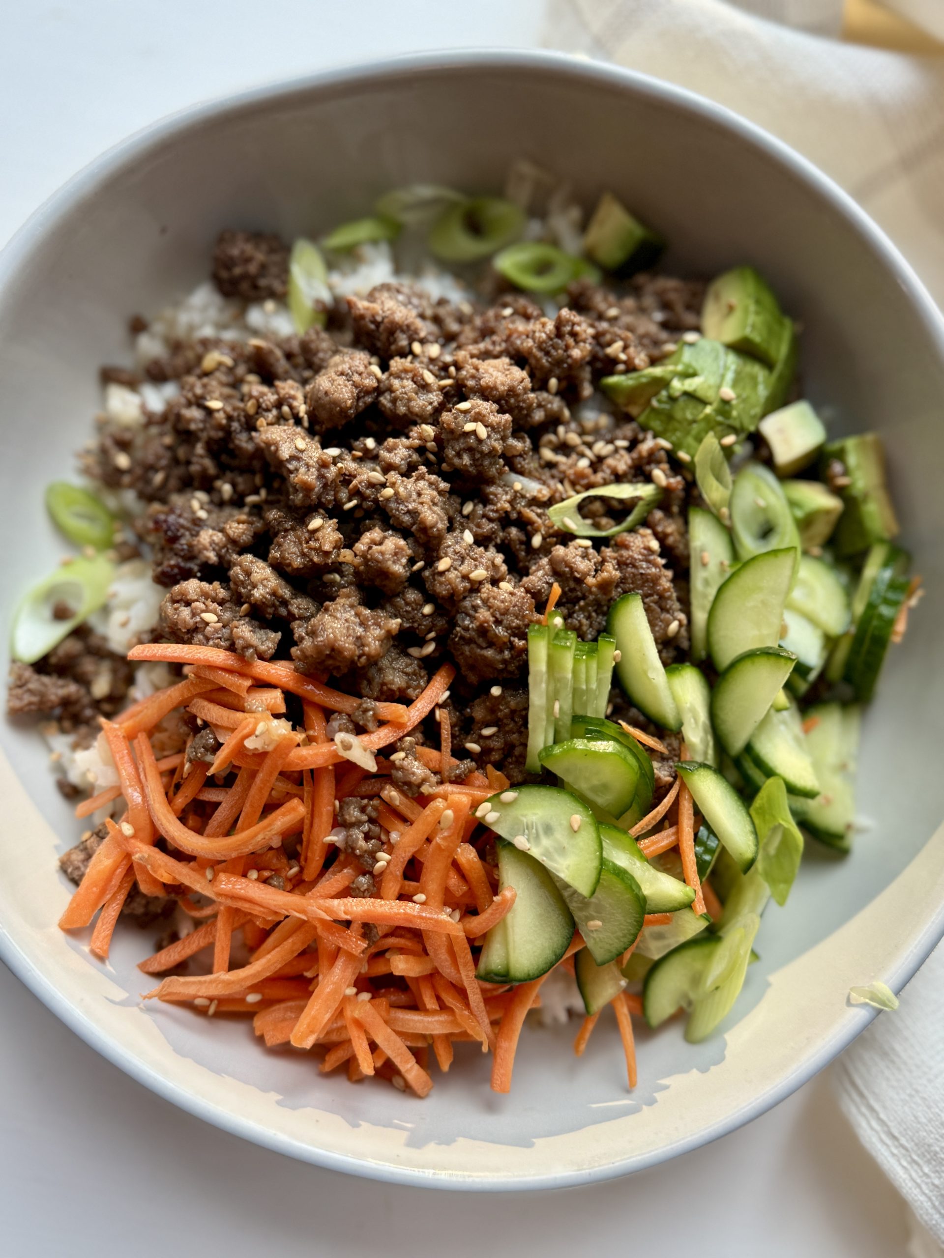 A bowl filled with ground meat, sliced cucumbers, shredded carrots, and chopped green onions, served over rice.