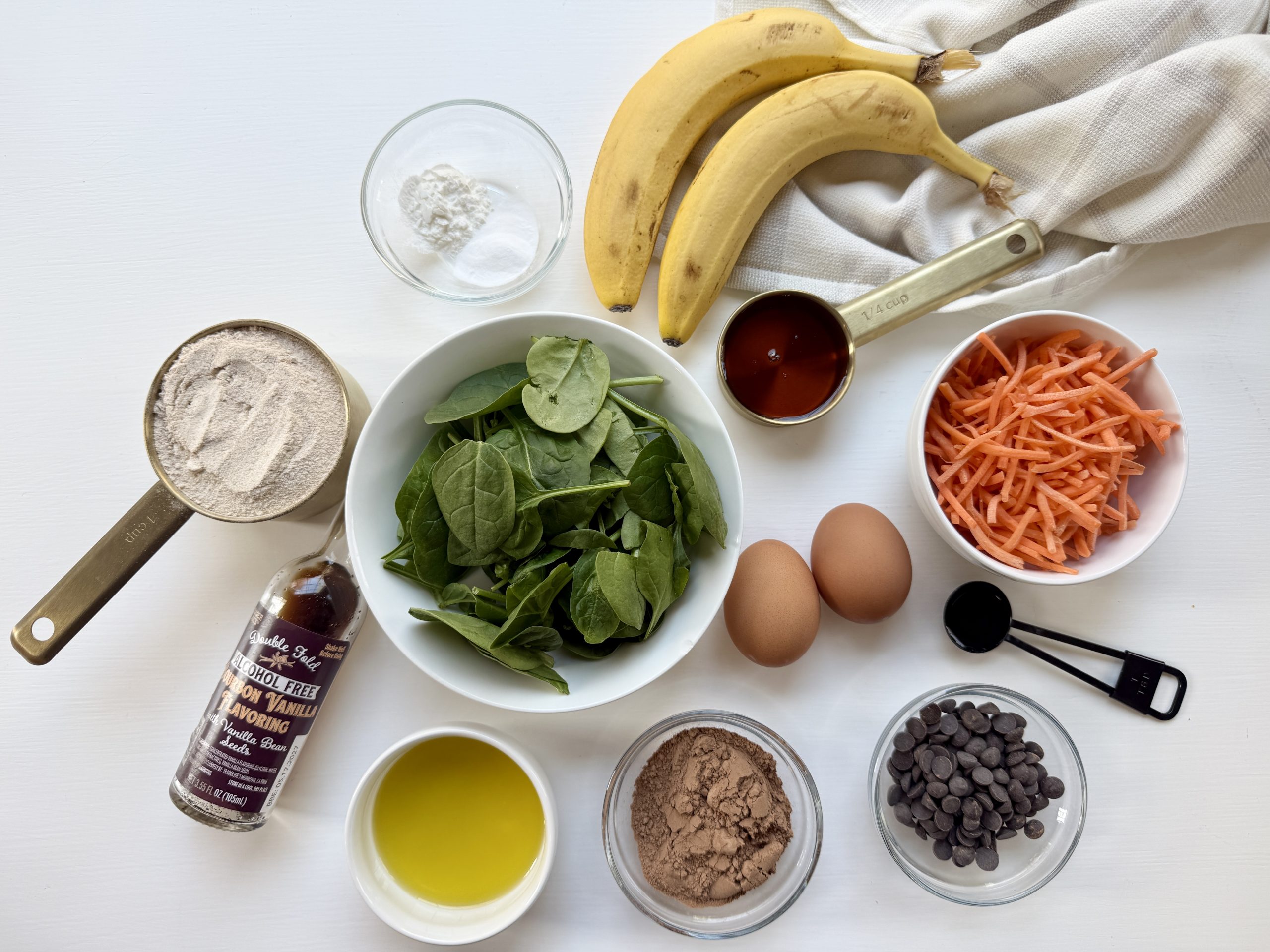 Various Chocolate Muffin ingredients, arranged in bowls on a white surface, including bananas, spinach, carrots, eggs, cocoa powder, flour, maple syrup, olive oil, and chocolate chips.