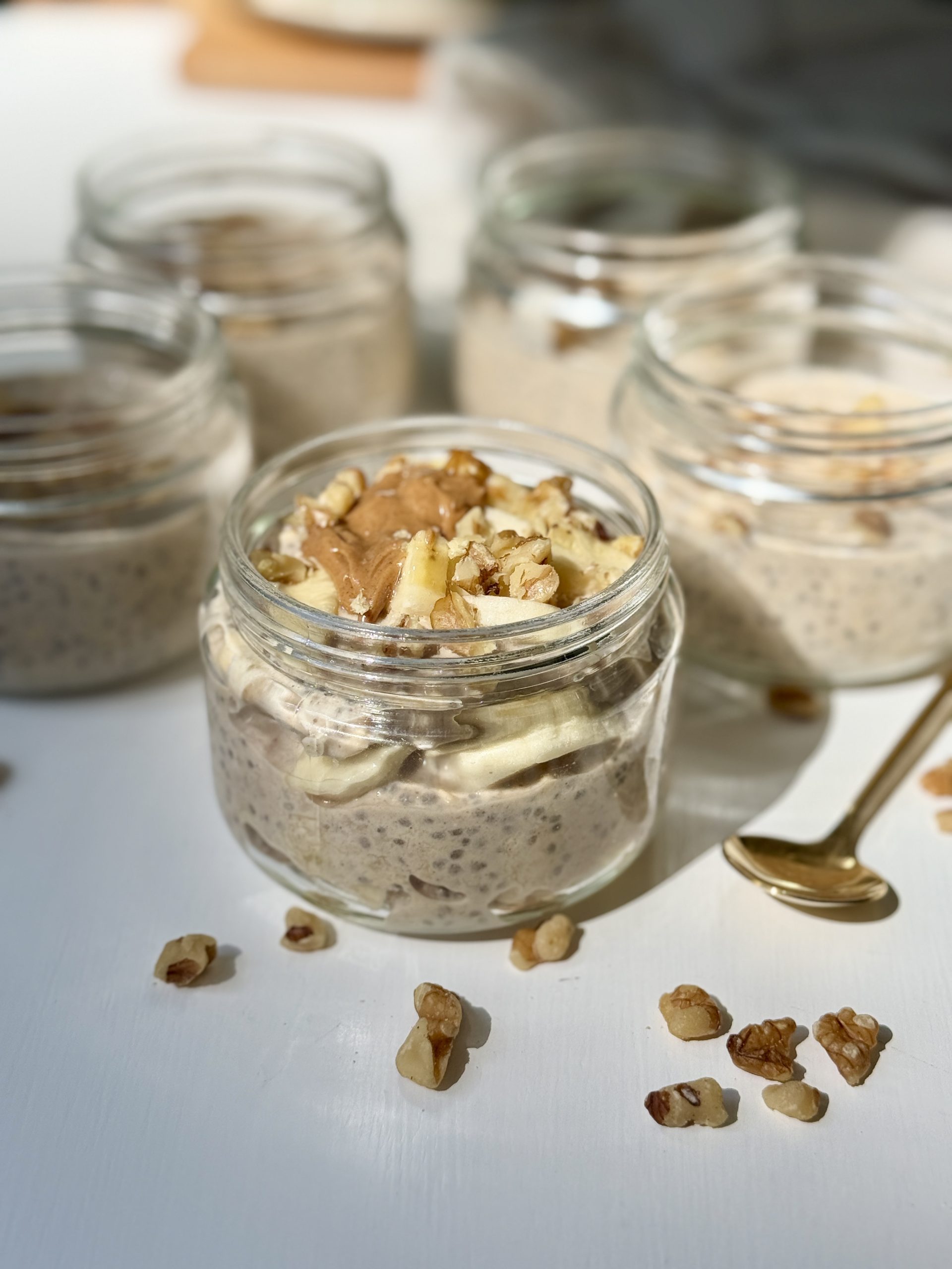Four glass jars filled with chia pudding are on a white surface. The front jar is topped with banana slices, walnuts, and nut butter, with a gold spoon and scattered walnut pieces nearby.