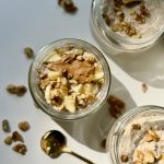Overhead view of three jars filled with creamy chia pudding topped with chopped walnuts and a dollop of nut butter. A gold spoon and scattered walnut pieces are on the white surface. Sunlight casts soft shadows.
