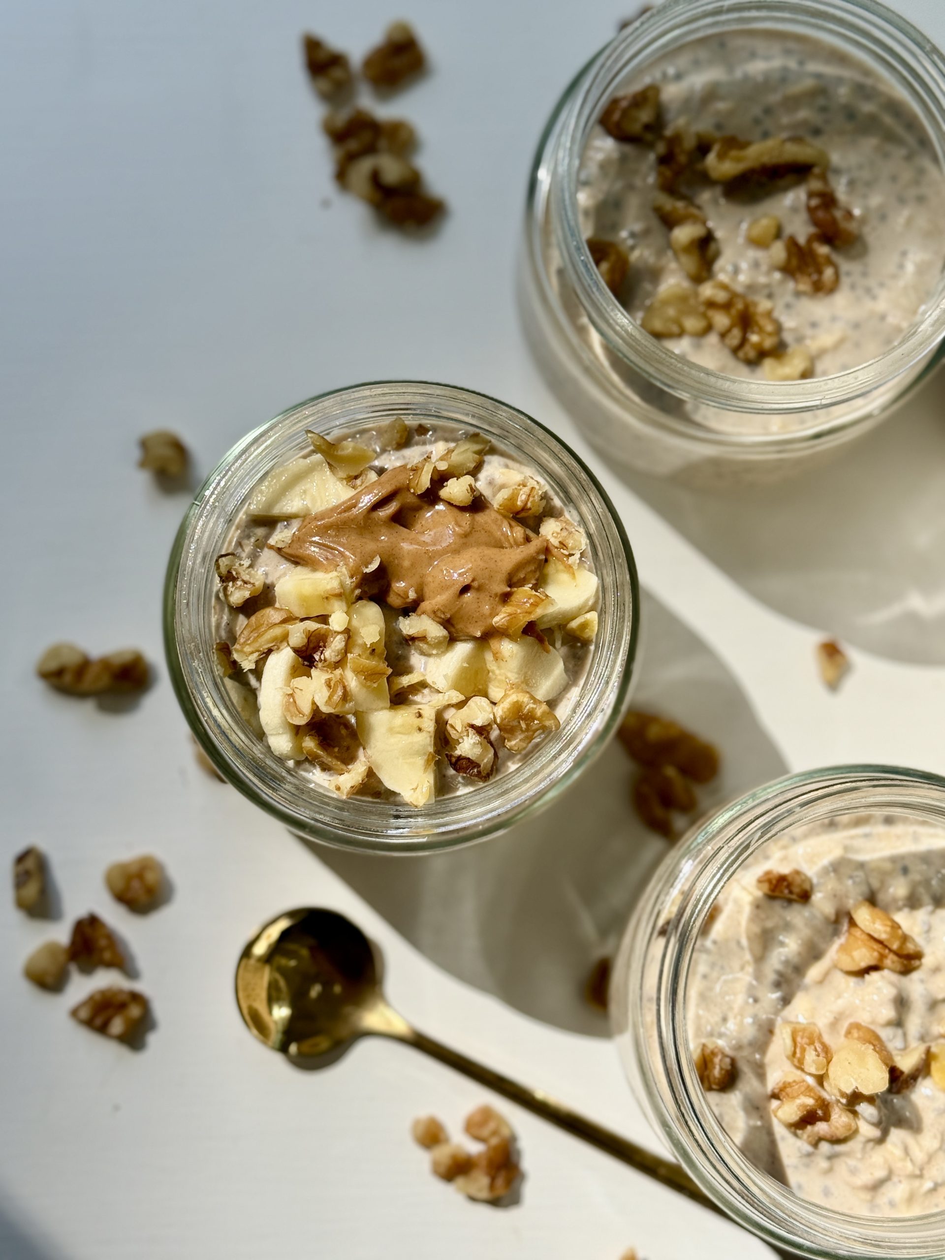Overhead view of three jars filled with creamy chia pudding topped with chopped walnuts and a dollop of nut butter. A gold spoon and scattered walnut pieces are on the white surface. Sunlight casts soft shadows.