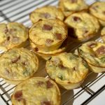 Mini egg bites with visible chunks of ham and vegetables rest on a wire cooling rack, bathed in bright sunlight on a white surface.