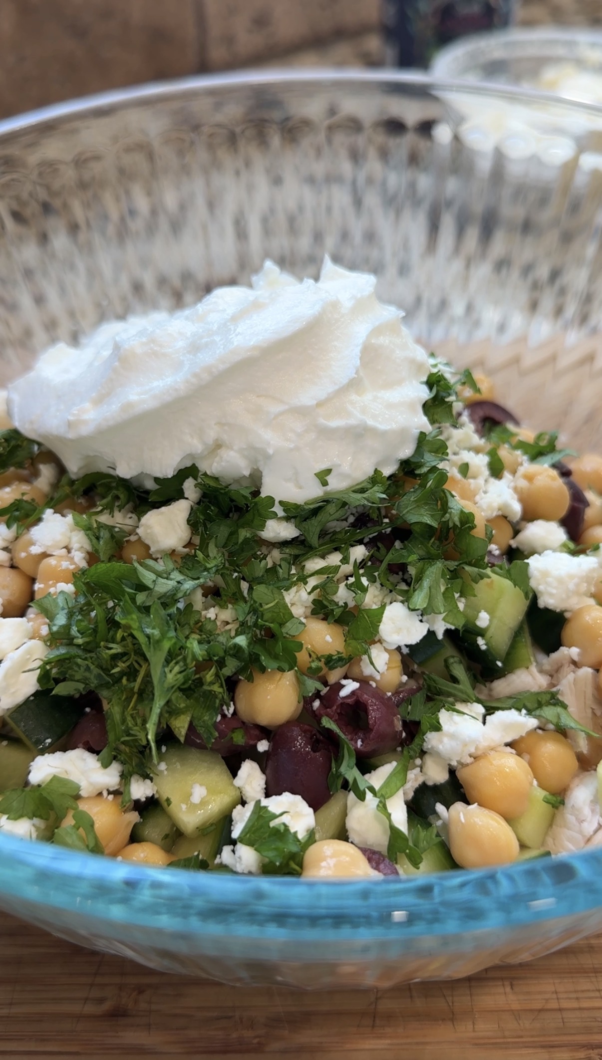 A close-up of a fresh salad with chickpeas, cucumbers, chopped parsley, kalamata olives, and crumbled feta cheese, topped with a generous dollop of whipped cream or Greek yogurt in a clear glass bowl.