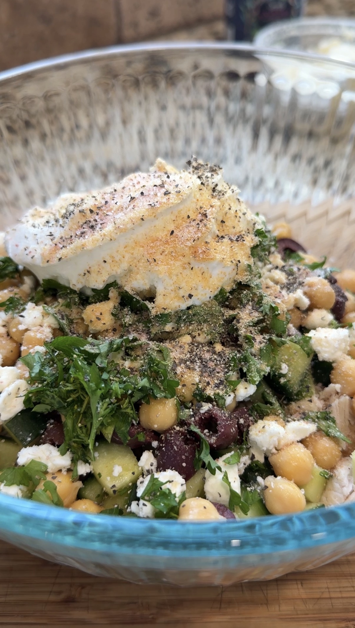 A glass bowl filled with a salad of chickpeas, chopped parsley, diced cucumber, and red kidney beans, topped with crumbled feta cheese, seasoning, and a dollop of creamy white dressing.
