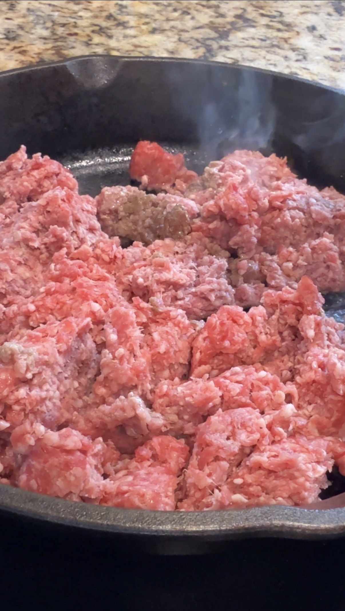 Raw ground beef is cooking in a skillet, with some areas beginning to brown while most of the meat remains pink and uncooked. Steam is visible, indicating the meat is being heated.