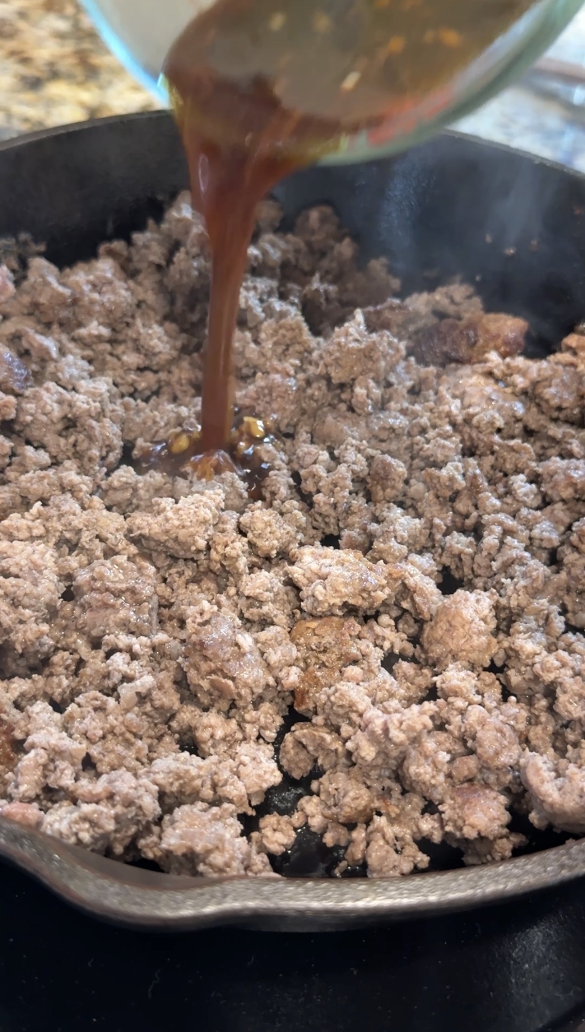 Cooked ground beef in a skillet with a brown sauce being poured over it, creating steam as the liquid touches the hot meat.