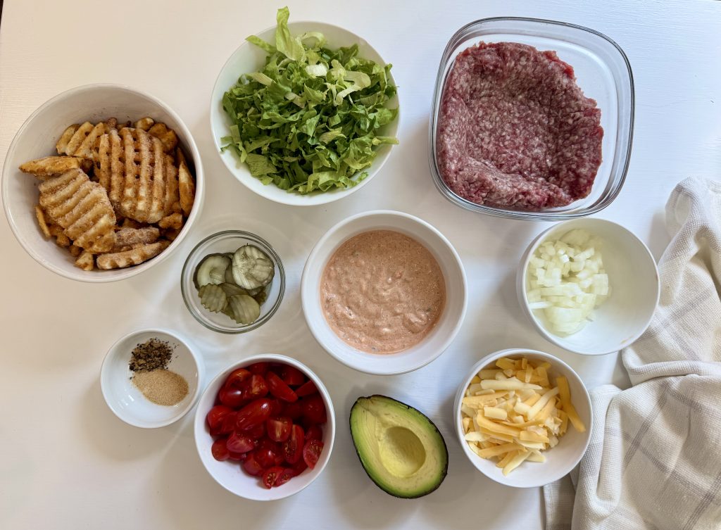 Various burger ingredients arranged in bowls on a white surface, including waffle fries, shredded lettuce, ground beef, pickles, sauce, diced onion, spices, halved grape tomatoes, half an avocado, and shredded cheese.