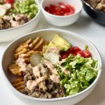 A bowl filled with waffle fries, ground beef, sliced pickles, avocado, cherry tomatoes, lettuce, and a drizzle of creamy sauce. Nearby are bowls of chopped lettuce, tomatoes, and ground beef.