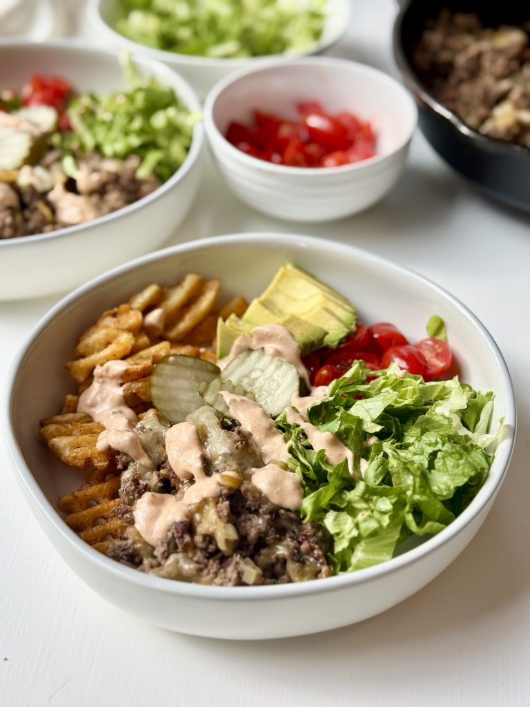 A bowl filled with waffle fries, ground beef, sliced pickles, avocado, cherry tomatoes, lettuce, and a drizzle of creamy sauce. Nearby are bowls of chopped lettuce, tomatoes, and ground beef.