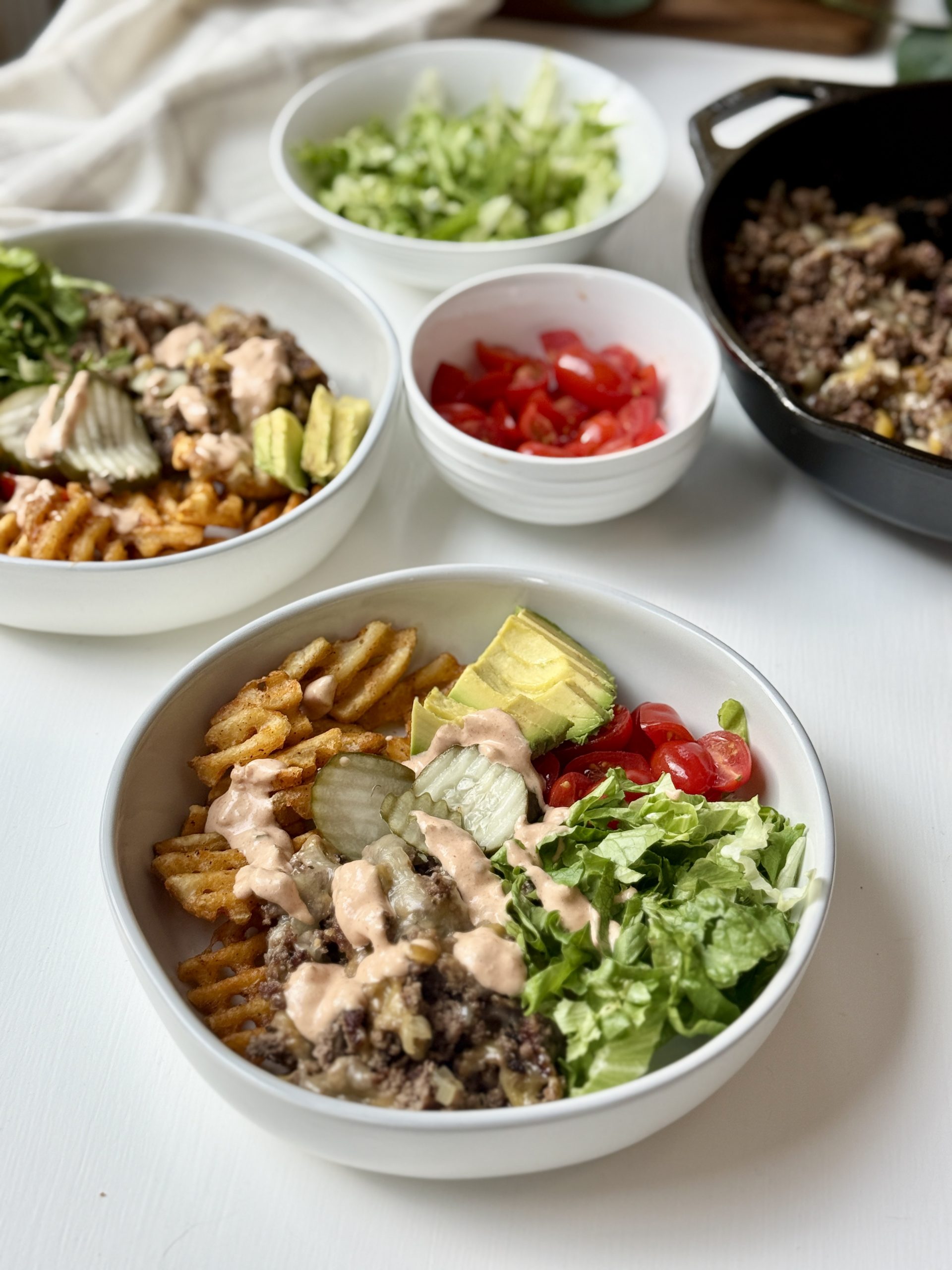 A bowl filled with waffle fries, ground beef, avocado, lettuce, tomato, pickles, and a creamy sauce. Other bowls with chopped lettuce, diced tomatoes, and more ground beef are nearby on a white table.