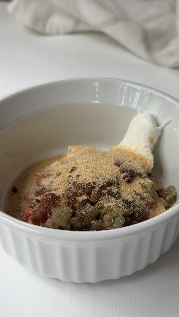 A white ramekin filled with dollops of white creamy sauce, green relish, and a blend of reddish and tan powdered seasonings, ready to be mixed.