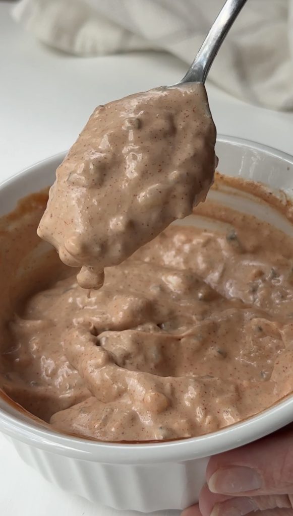 A close-up view of a thick, creamy, reddish-brown sauce with visible seasoning, being scooped from a white bowl with a spoon.
