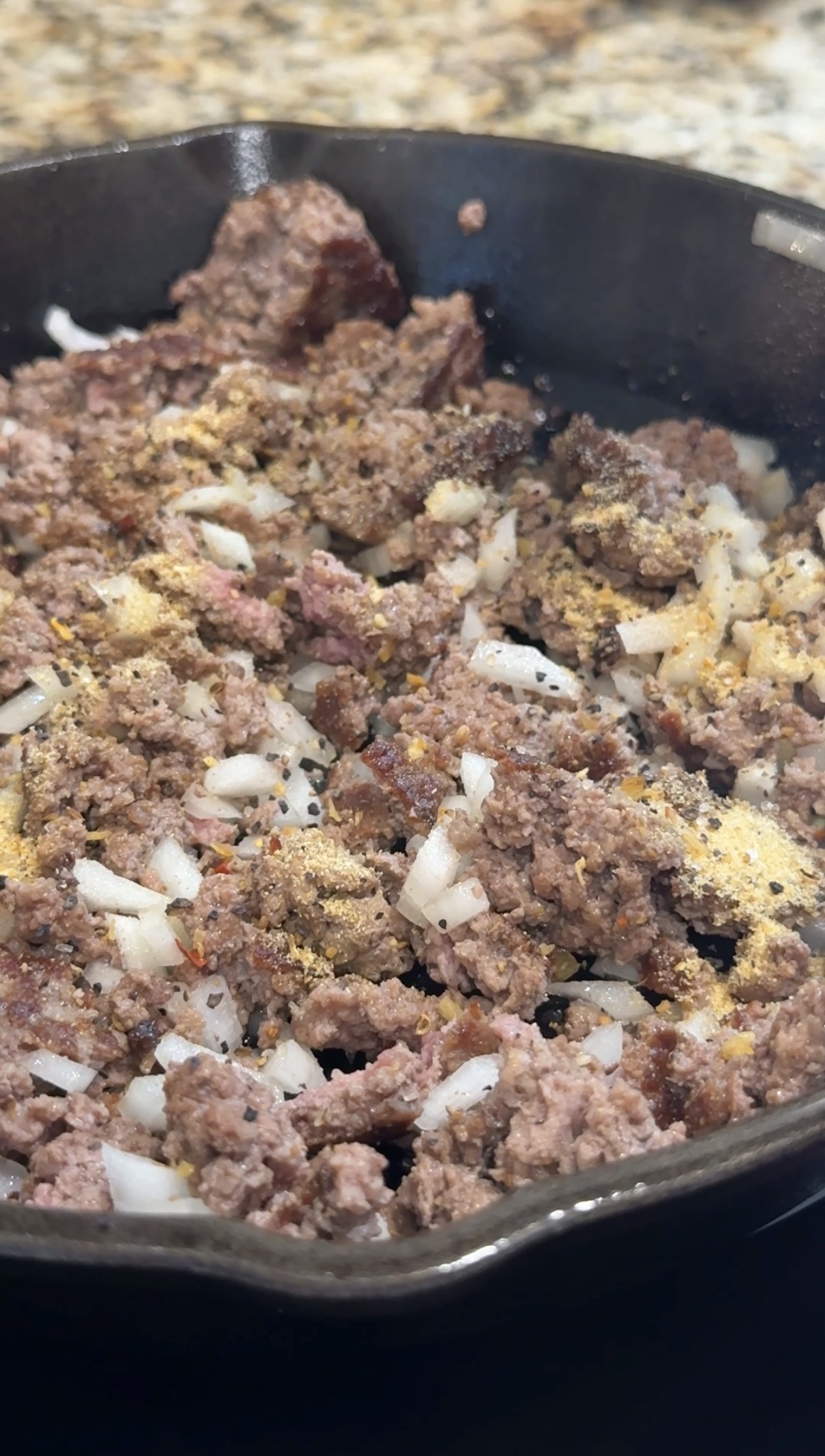 Ground beef and chopped onions cooking in a skillet, topped with various seasonings including dried herbs and powdered spices.