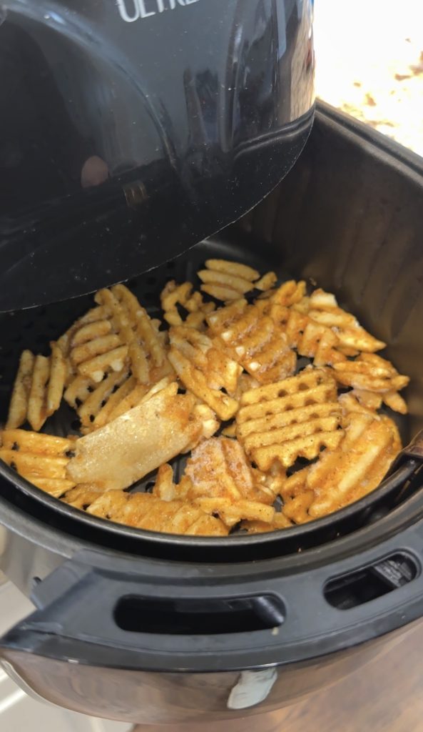Waffle fries and a piece of chicken are cooking inside an open air fryer basket. The fries are golden and crispy, and the air fryer lid is partially lifted.