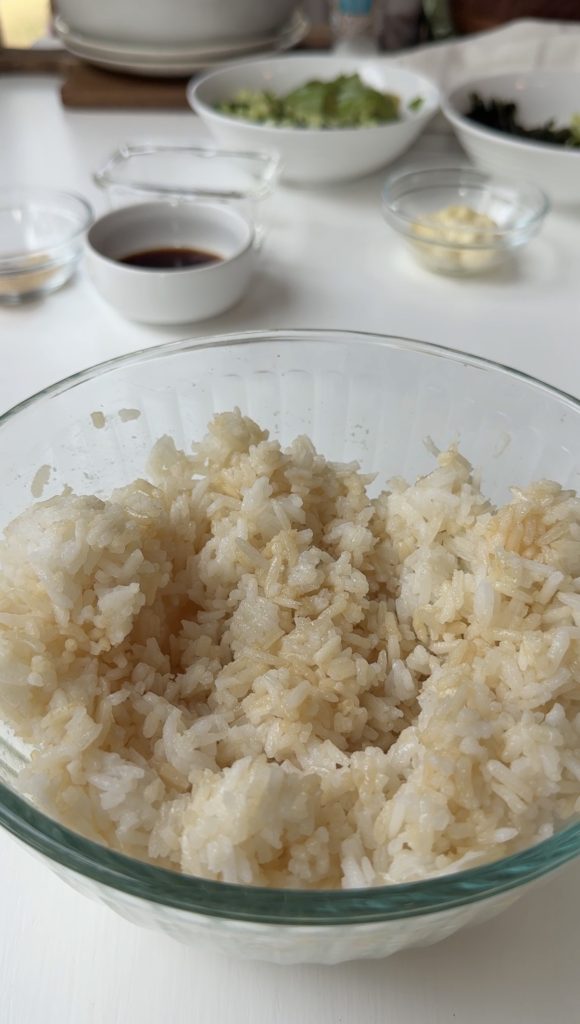 A clear glass bowl filled with steamed white rice drizzled with soy sauce sits on a white table, with small bowls of condiments and ingredients blurred in the background.