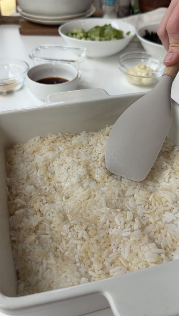A person uses a spatula to spread cooked rice in a white square dish. Bowls of soy sauce, water, and other ingredients are visible in the background on a white surface.