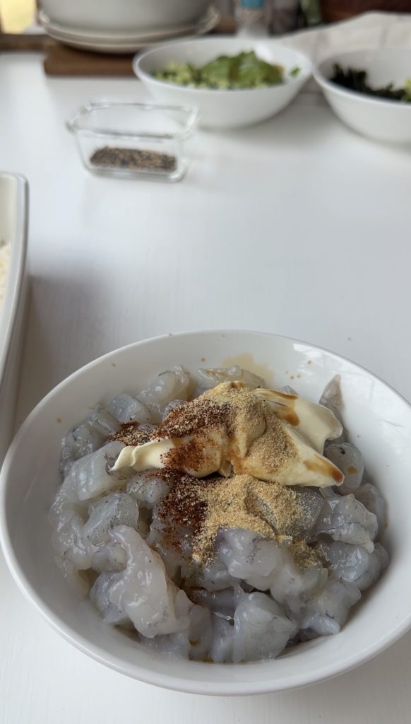 A white bowl filled with raw, chopped shrimp topped with spices, mayonnaise, and seasoning. Other bowls with ingredients are blurred in the background on a white table.