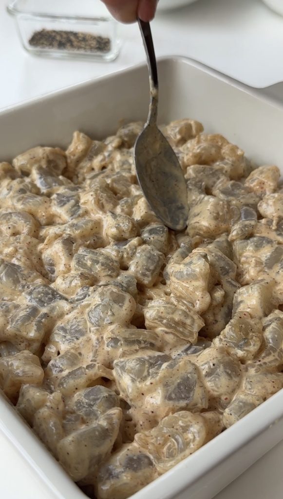 A close-up of a white dish filled with creamy, diced pieces of food being stirred with a spoon. The mixture appears to be coated in a thick, seasoned sauce.