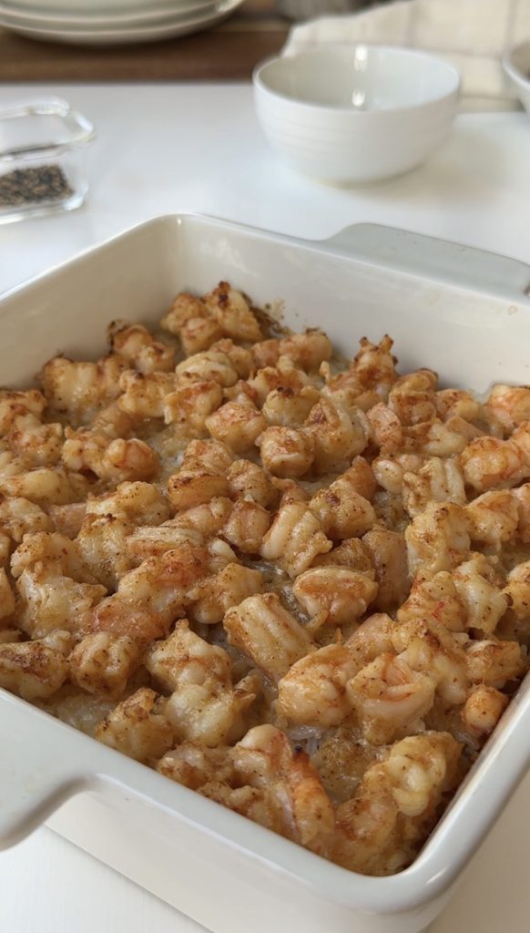 A white ceramic baking dish filled with cooked, seasoned shrimp pieces sits on a white table, with bowls and a dish of pepper visible in the background.