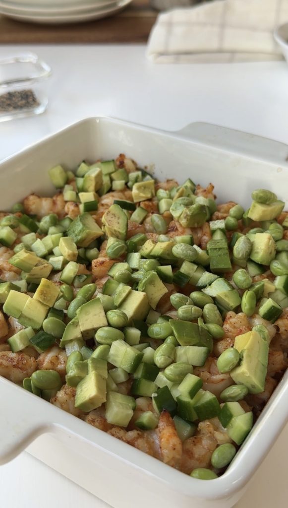 A white square dish filled with a baked casserole topped with diced avocado, edamame, and cucumber, sitting on a white table. Plates and a small glass dish are visible in the background.