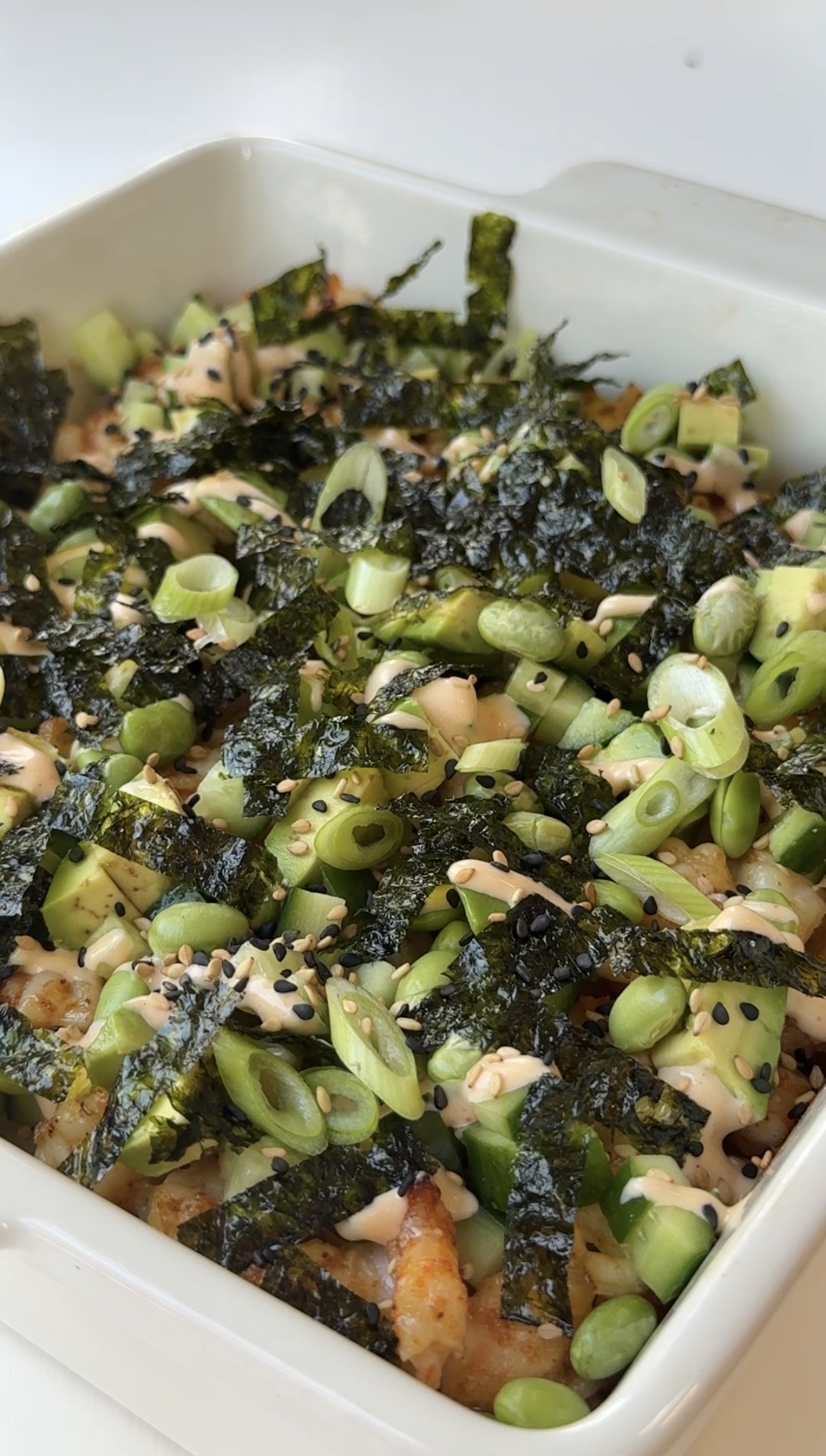 A close-up of a baked sushi casserole topped with sliced green onions, shredded seaweed, sesame seeds, and a drizzle of sauce in a white square dish.