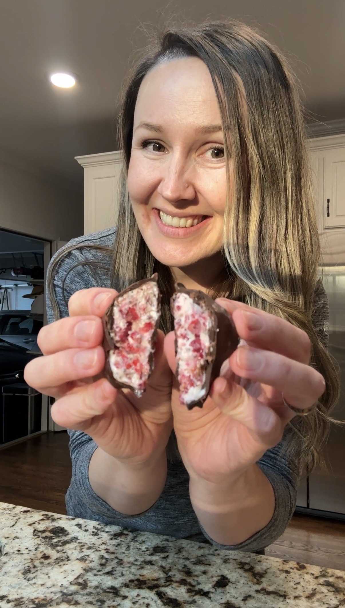 A smiling woman with long hair holds a chocolate-covered treat split in half, revealing a creamy white and pink filling. She is standing in a kitchen with white cabinets in the background.