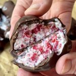 A hand holds a chocolate-coated treat broken in half, revealing a creamy filling mixed with bright red berry pieces. The background shows more of the same treats on a brown paper surface.