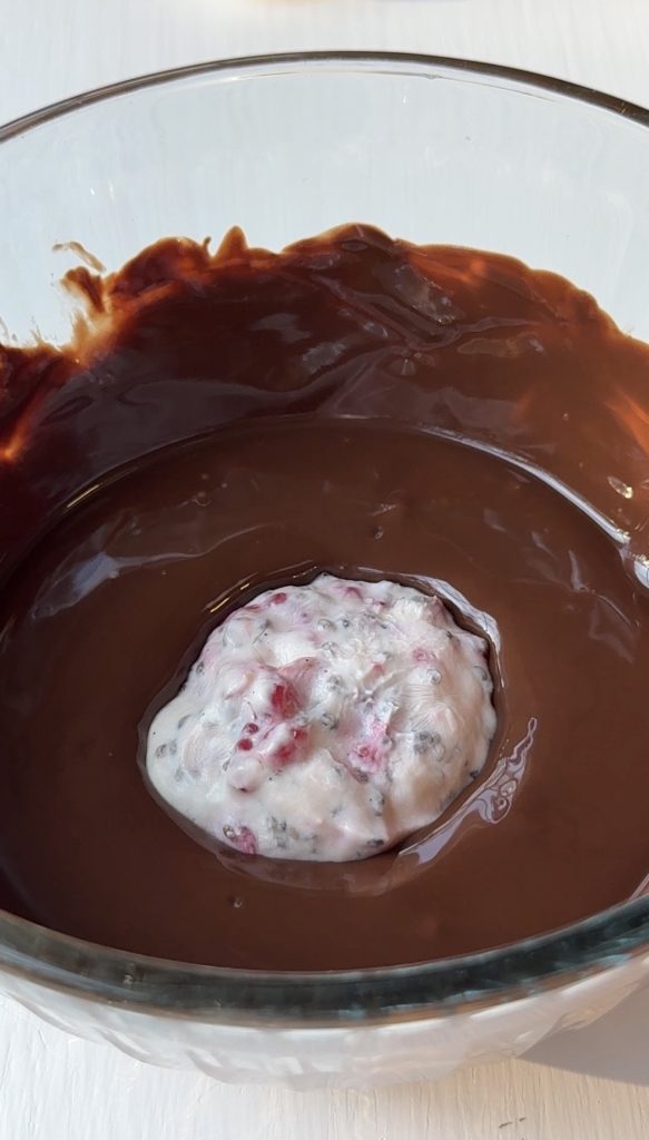 A round ball of creamy white mixture with pink and dark specks is partially submerged in a glass bowl filled with melted chocolate. Chocolate coats the sides of the bowl.