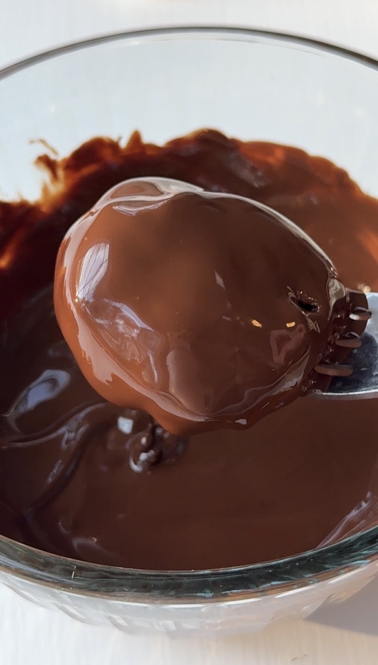 A close-up of a fork holding a round treat coated in melted chocolate above a bowl filled with more melted chocolate.