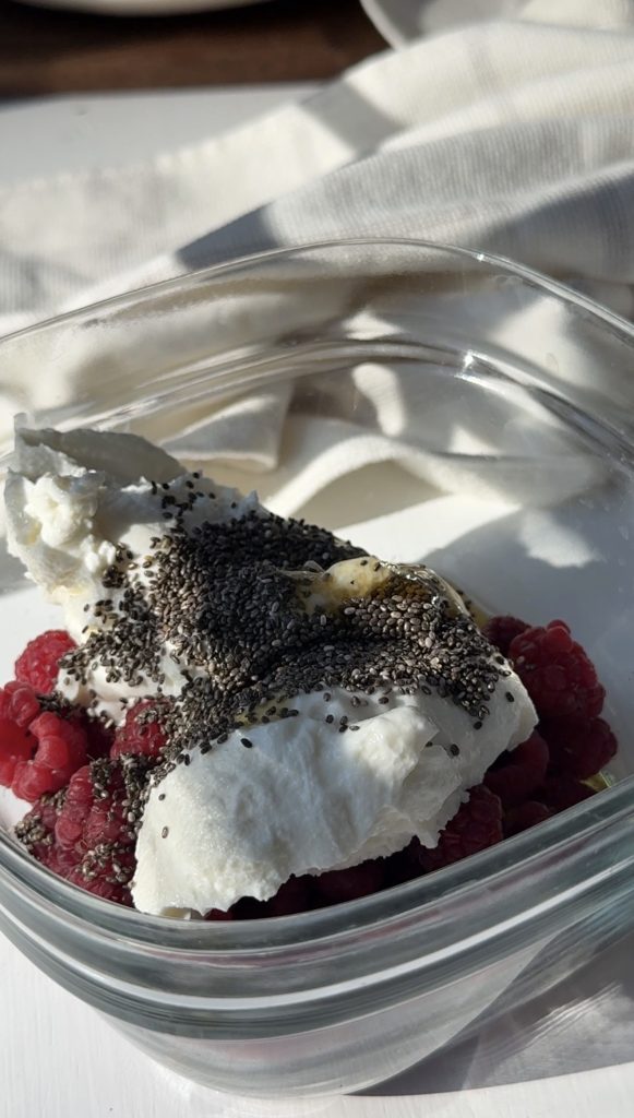 A glass container filled with fresh raspberries, topped with dollops of Greek yogurt, chia seeds, and a drizzle of honey, placed on a white surface with soft natural light.