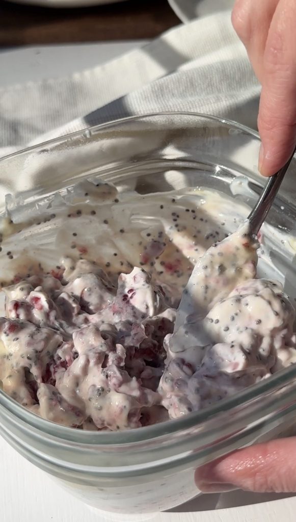 A hand holds a spoon in a glass bowl filled with a creamy mixture containing visible chia seeds and pieces of berries or fruit. The sunlight highlights the texture and ingredients of the mixture.