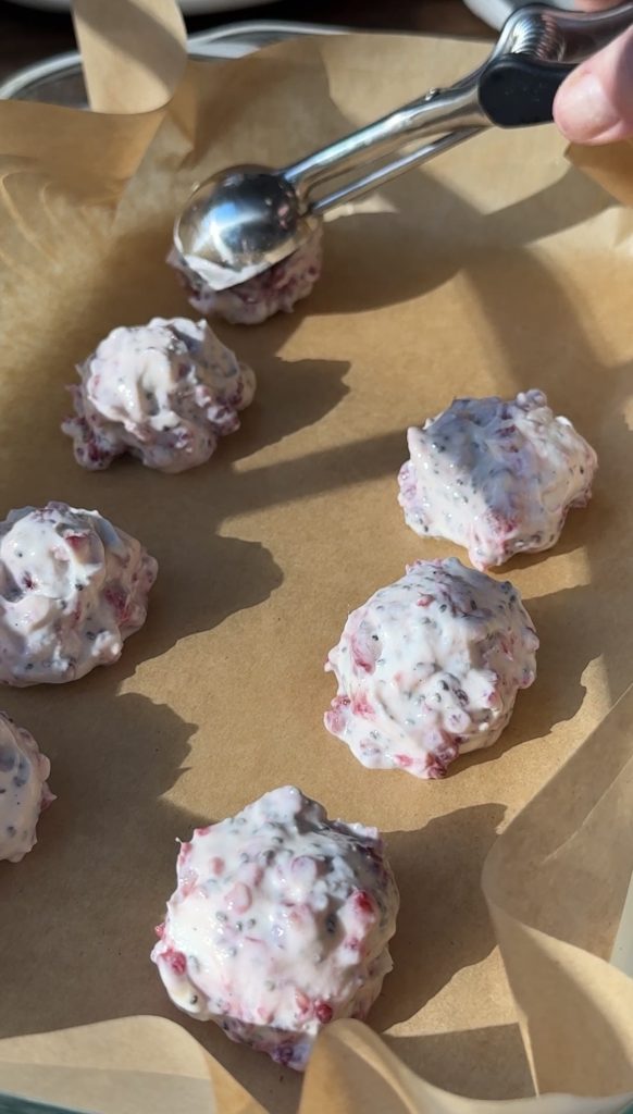 Scoops of a creamy mixture with visible purple and pink berry pieces are placed on a sheet of parchment paper. A metal scoop is seen in the background, used for portioning the mixture.